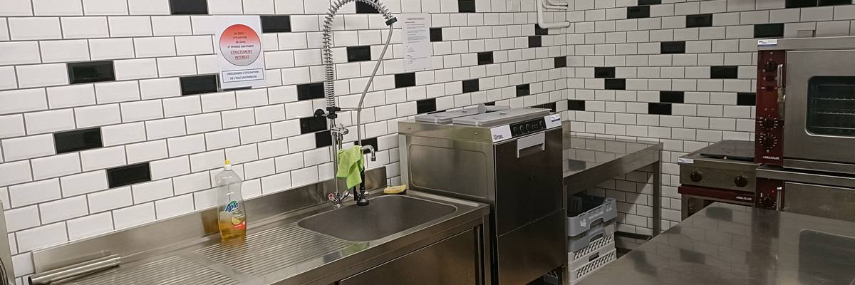 Cuisine professionnelle avec évier, lave-vaisselle, plan de travail en acier inoxydable, four et chauffe-eau mural.