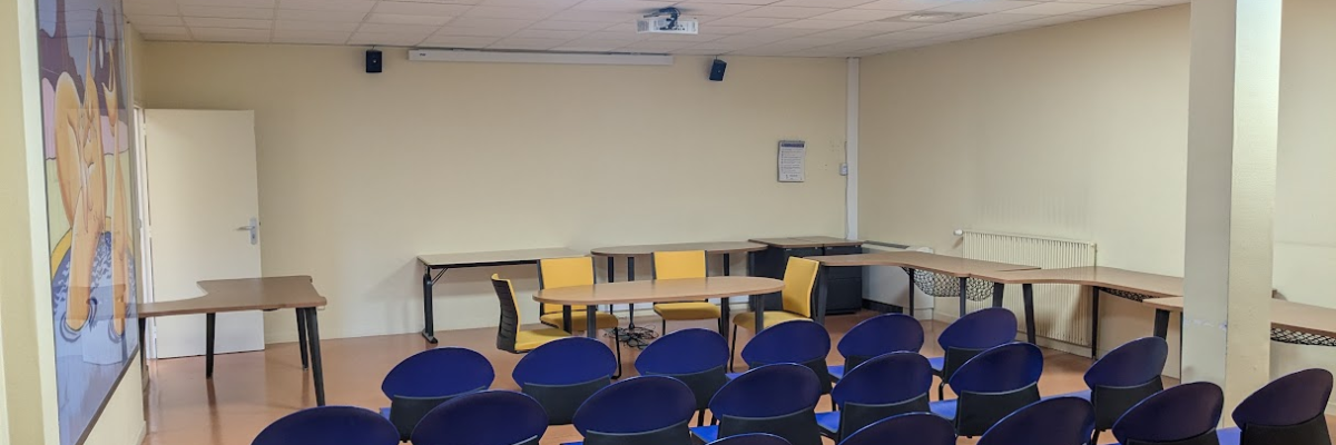 Salle avec rangées de chaises bleues, tables, projecteur, écran et tableau.