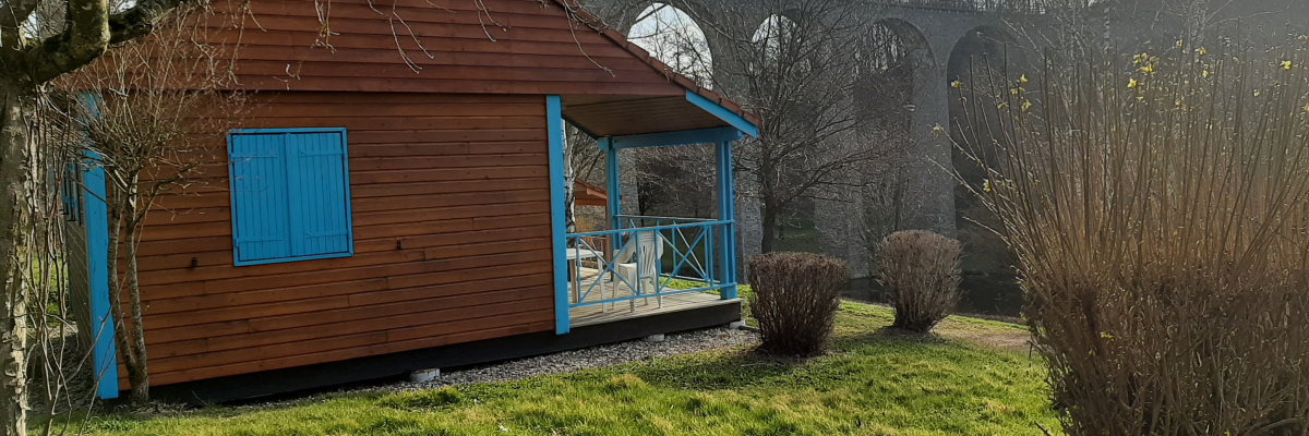 Pavillon en bois avec fenêtres bleues et terrasse, pont en pierre en arrière-plan, jardin avec herbe et buissons.