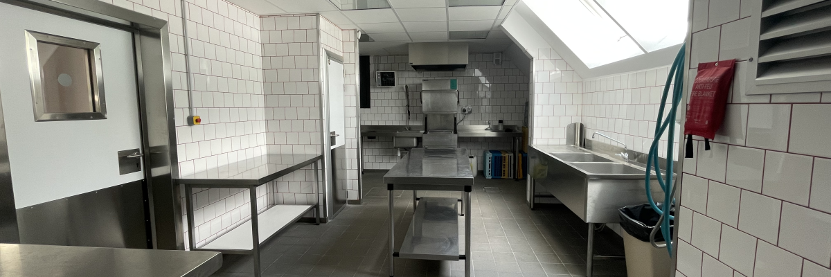 Cuisine professionnelle avec surfaces métalliques, éviers, robinetterie, tables de travail, étagères et carrelage mural blanc.