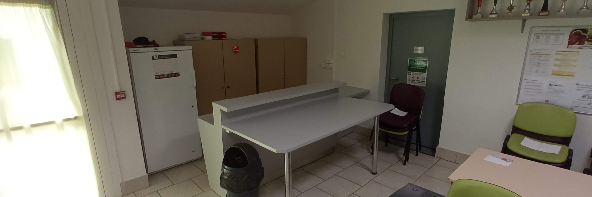 Salle avec table, chaises, armoires, frigo, poubelle, trophées et affichage.