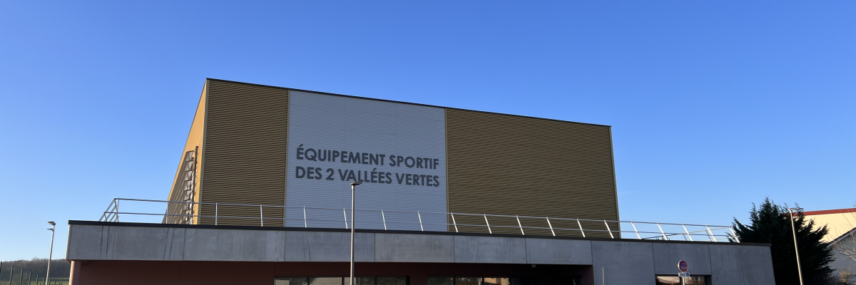 Bâtiment avec façade en bois et verre, parking et panneau "Équipement sportif des 2 vallées vertes".