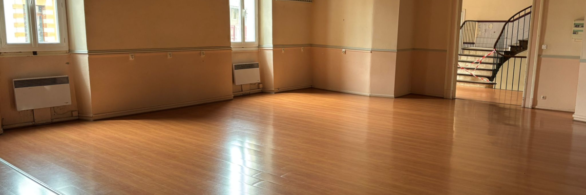 Salle vide avec parquet, radiateurs, fenêtres et escalier.