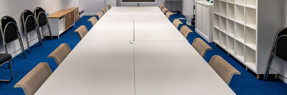 Salle de réunion blanche avec table, chaises, écran, étagères et meuble de rangement.