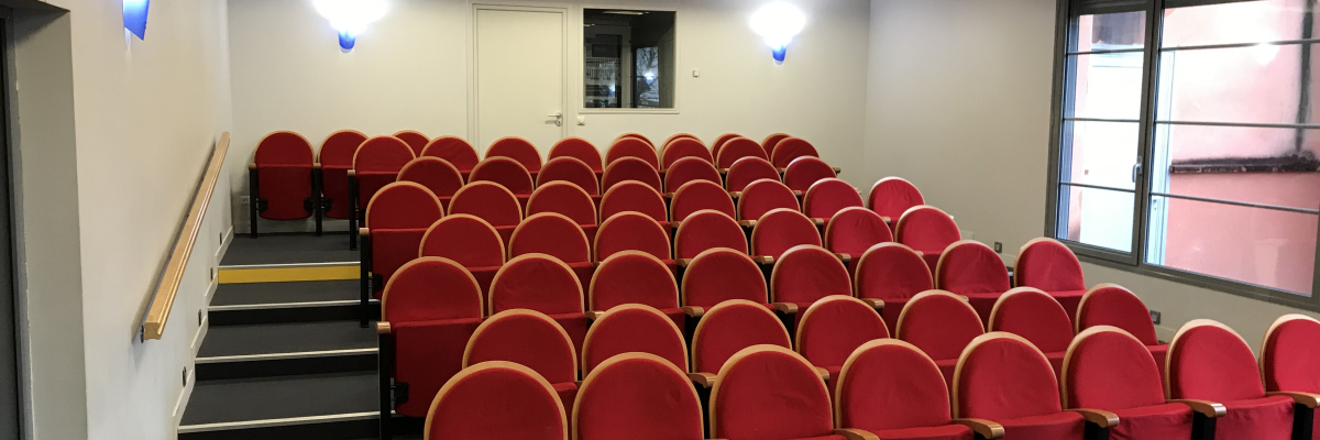 Salle avec rangées de chaises rouges, vidéoprojecteur, éclairage et fenêtres.
