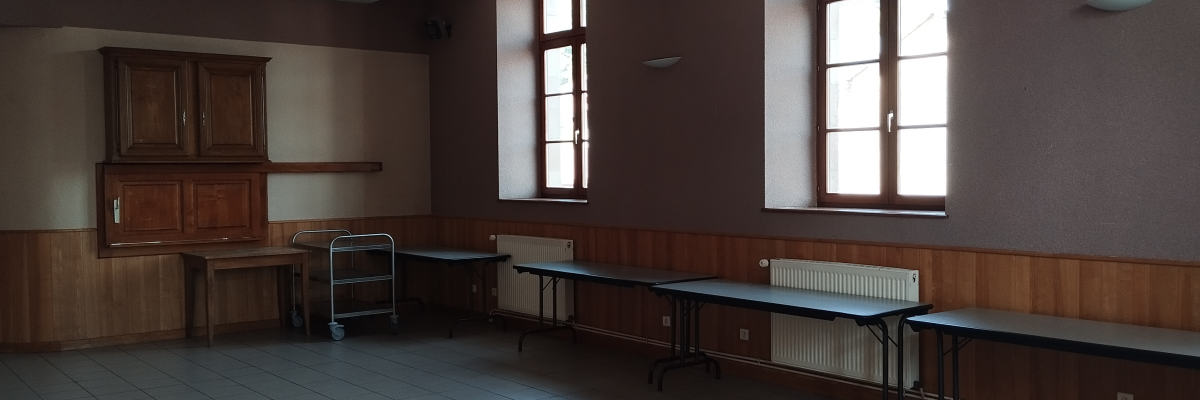 Salle avec tables, chaises, chariot, armoire, radiateurs et fenêtres.