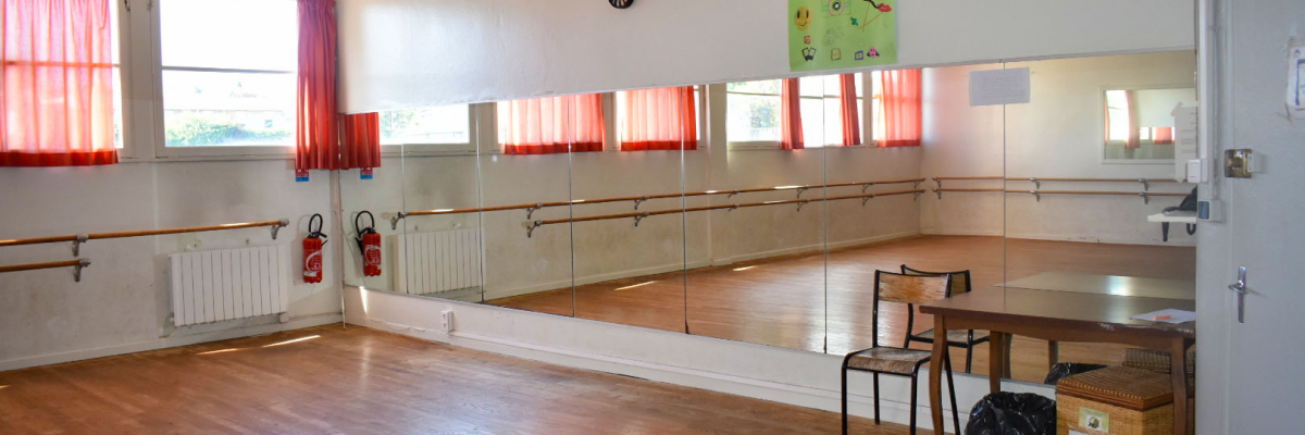 Salle de danse avec miroirs, barres, radiateurs, table, chaise, extincteur et horloge.