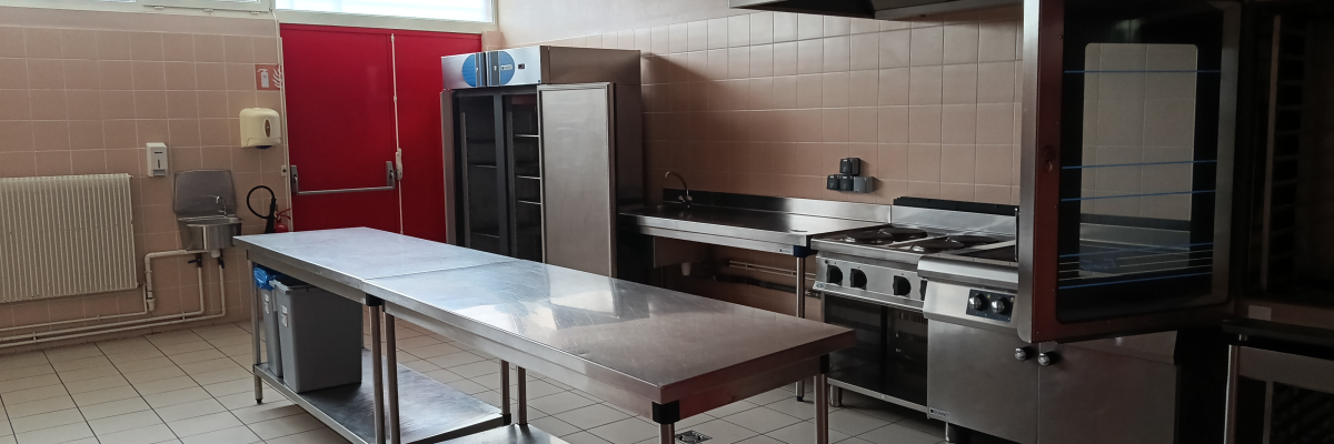 Cuisine avec tables en acier inoxydable, fours, plaques de cuisson, hotte, réfrigérateur et évier.