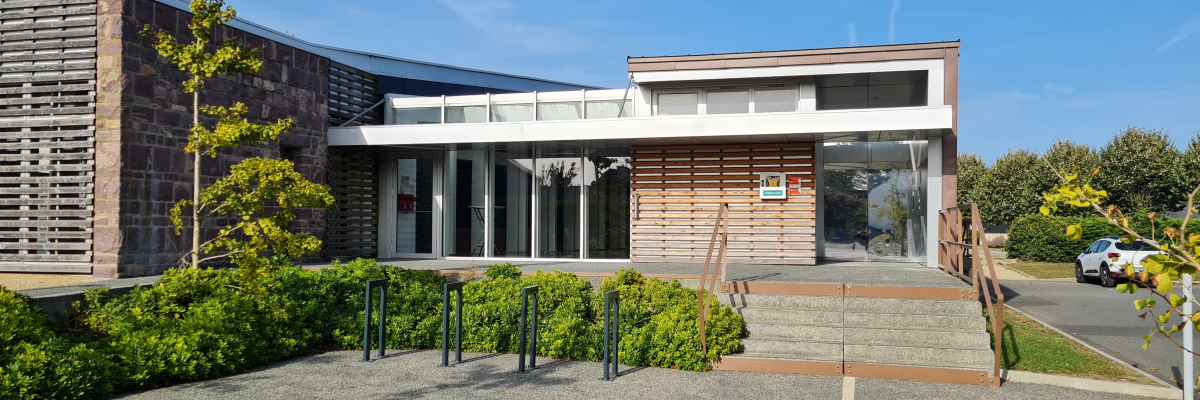 Bâtiment avec entrée vitrée, arbre, buissons et parking.