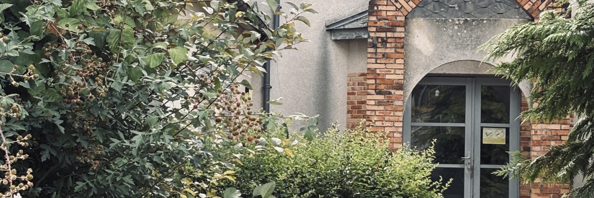 Maison avec toit en ardoise, briques apparentes, porte d'entrée et végétation environnante.