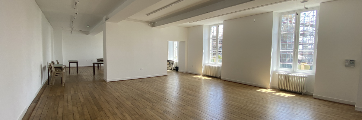 Grande salle vide avec plancher en bois, éclairage encastré, fenêtres et radiateur.
