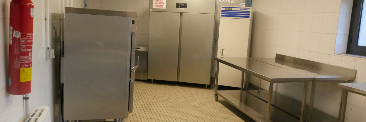 Cuisine industrielle avec plans de travail, réfrigérateurs, étagères et extincteur.