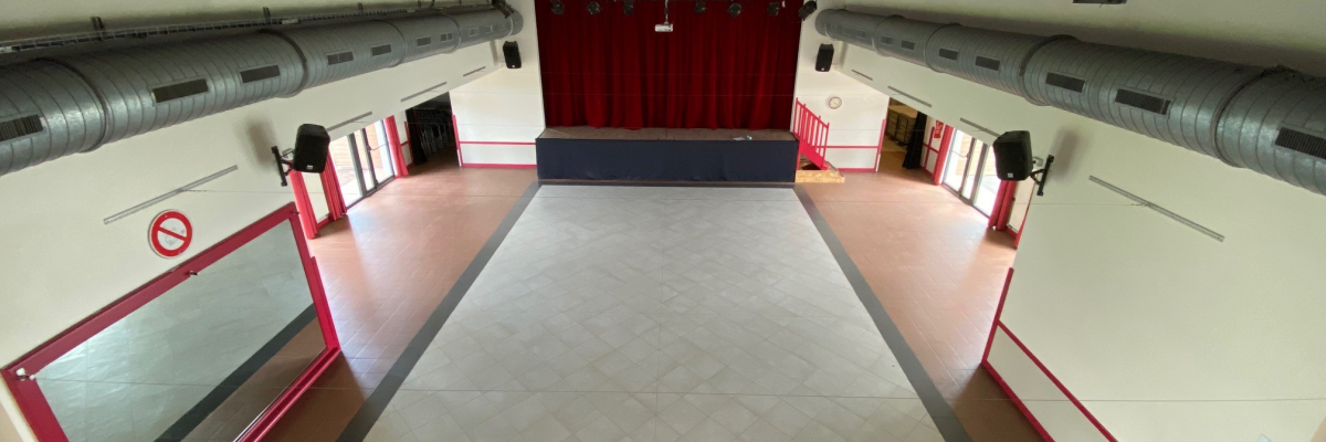 Salle avec scène, rideaux rouges, murs blancs, sol carrelé, conduits aériens, portes et spots.