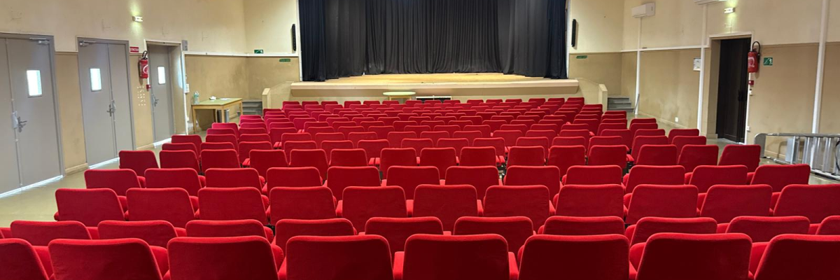 Salle avec rangées de sièges rouges, scène, rideau, éclairage et portes.
