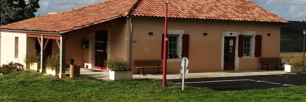 Maison basse avec toiture en tuiles rouges, portique, lampadaire et bancs près d'une aire de stationnement.