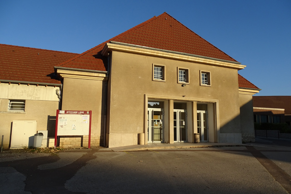 Bâtiment avec toit rouge, entrée principale en verre, un panneau de stationnement et une poubelle visible.