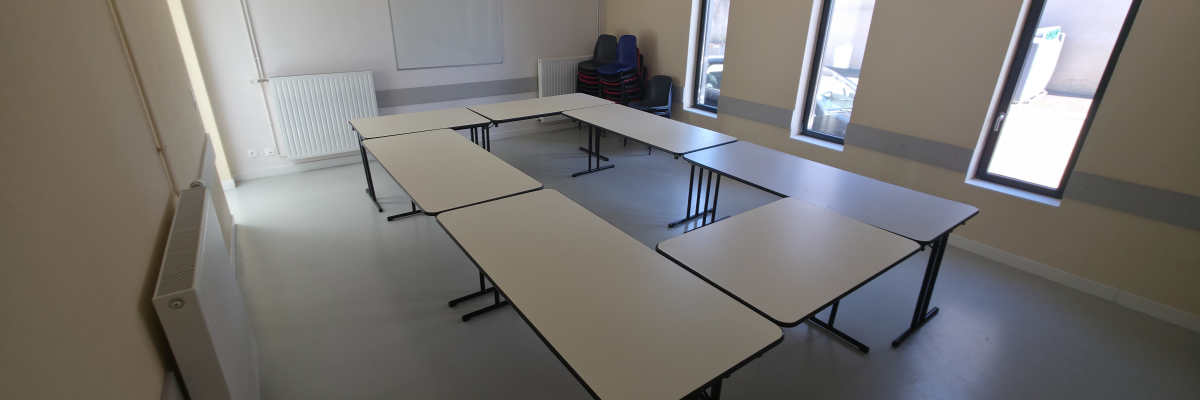 Salle avec tables, chaises, tableau blanc, radiateur et fenêtres.