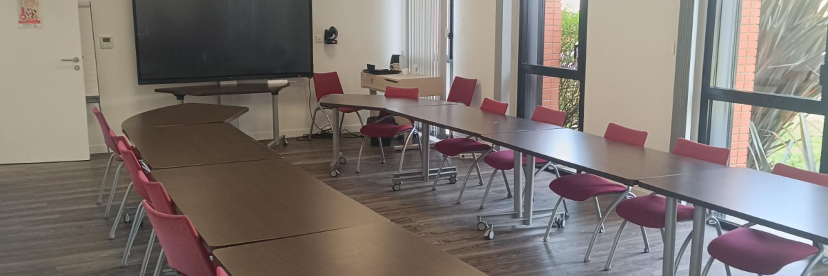 Salle de réunion avec tables, chaises, écran et fenêtres.