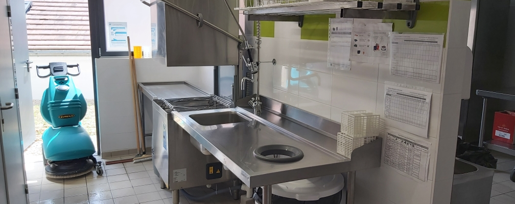 Laboratoire avec paillasse, évier, lave-vaisselle, étagère, documents, balai et seau.