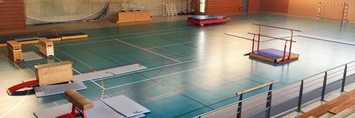 Salle de sport avec tapis, cheval d'arçons, barres parallèles, barre fixe, agrès, trampoline et gradins.