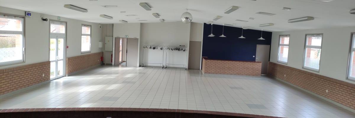 Salle vide avec murs partiels en brique, sol carrelé, luminaires suspendus, porte, fenêtres et tableau.