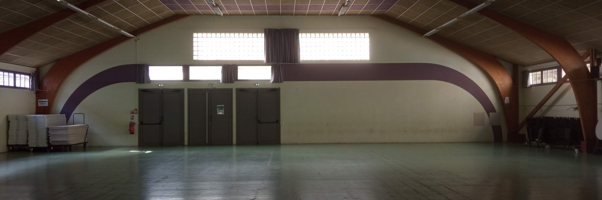 Gymnase avec sols en bois, murs courbés, grandes fenêtres, portes et quelques chaises.