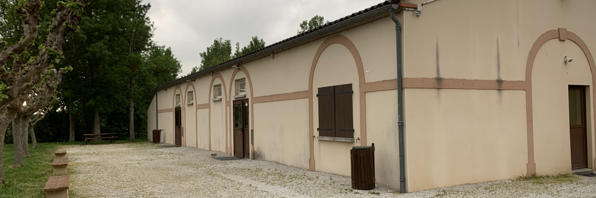 Bâtiment rectangulaire avec murs beiges, portes et fenêtres sombres, gravier au sol, poubelle extérieure et arbre à gauche.