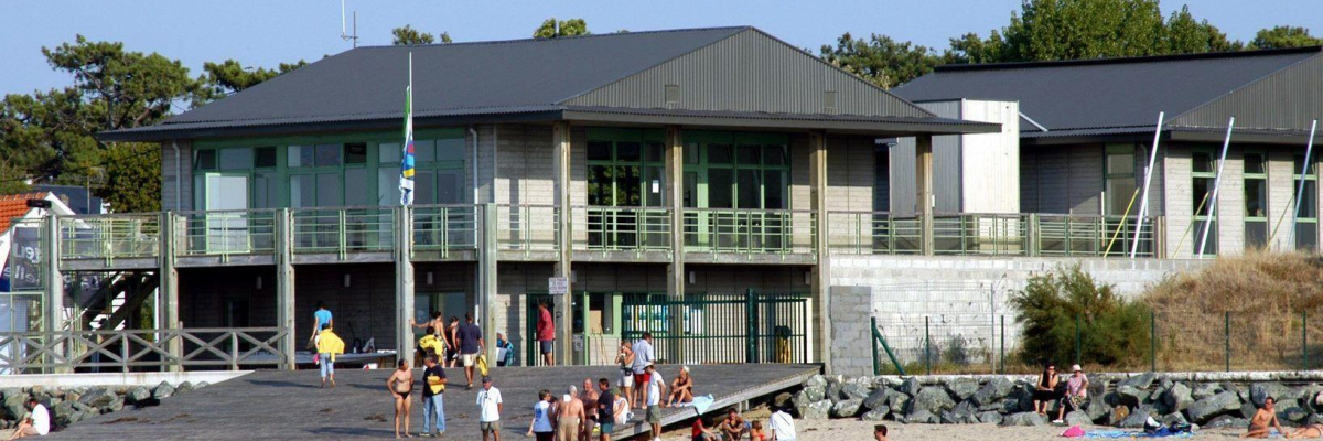Bâtiment moderne avec balcons, plage, gens, canoës, drapeau.