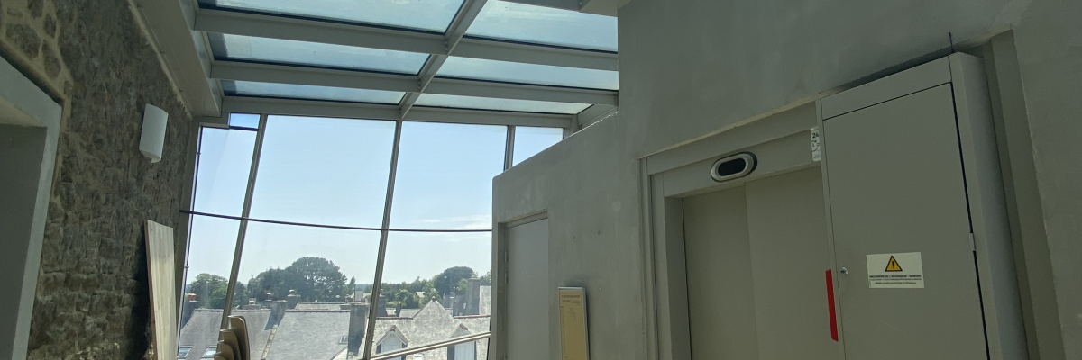 Couloir avec toit vitré, porte de sortie, chaise et mur en pierre.