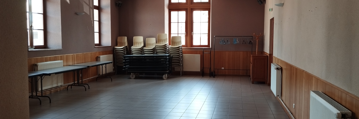 Salle vide avec tables, chaises empilées, radiateurs et tableau. Fenêtres et éclairage au plafond.