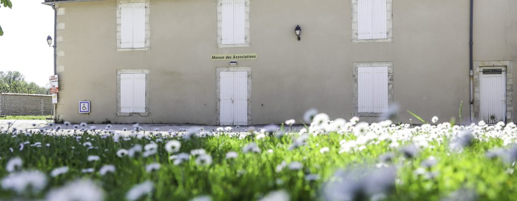 Maison avec murs beiges, six fenêtres fermées, deux portes principales, une enseigne "Maison de l'Association" et un lampadaire.