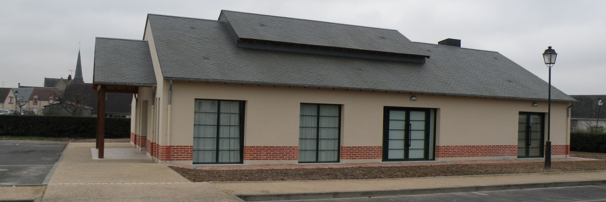 Bâtiment avec toit gris, murs beiges et briques rouges, grandes fenêtres, parking et réverbère.