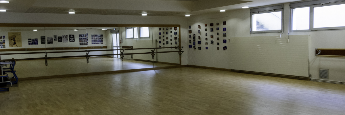 Salle avec barres de ballet, miroir, poubelle, et rangée de lumières au plafond.