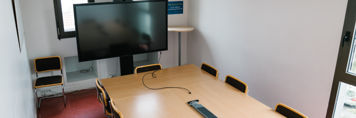 Salle avec table, chaises, écran, câbles et fenêtres.