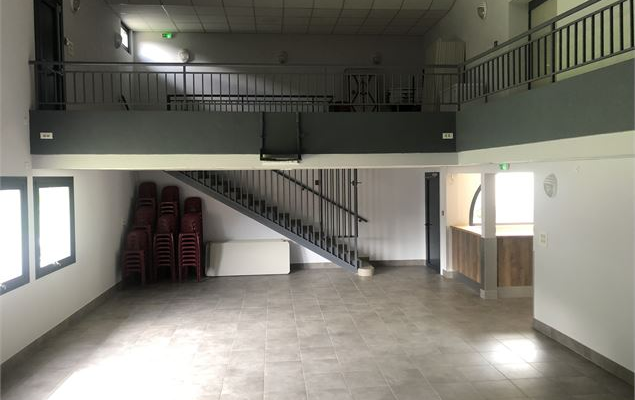 Salle vide avec escalier, mezzanine, portes et fenêtres. Chaises empilées près de l'escalier.