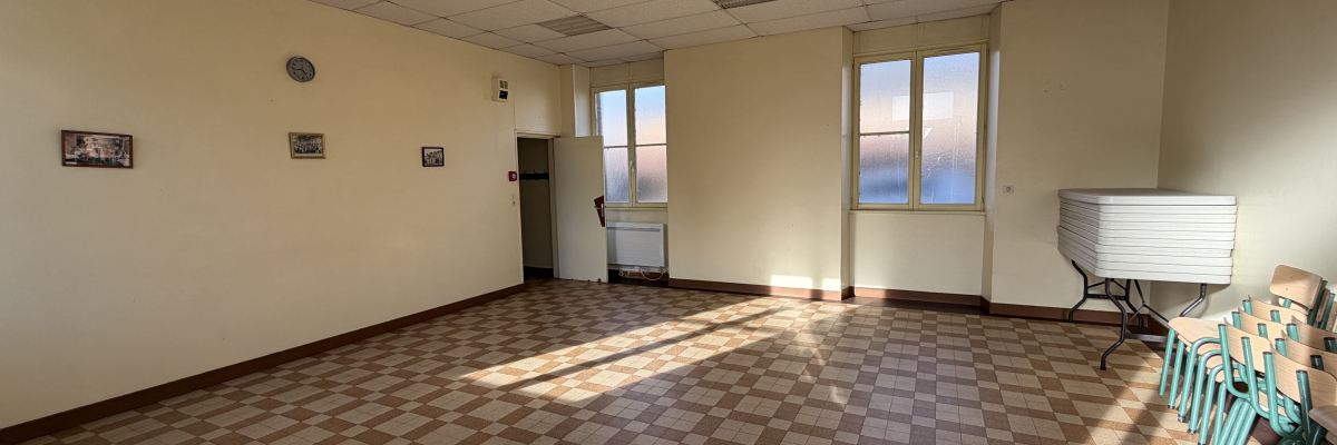 Pièce vide avec sol carrelé, plafond à dalles, fenêtres, radiateur, ventilateur et chaises empilées.