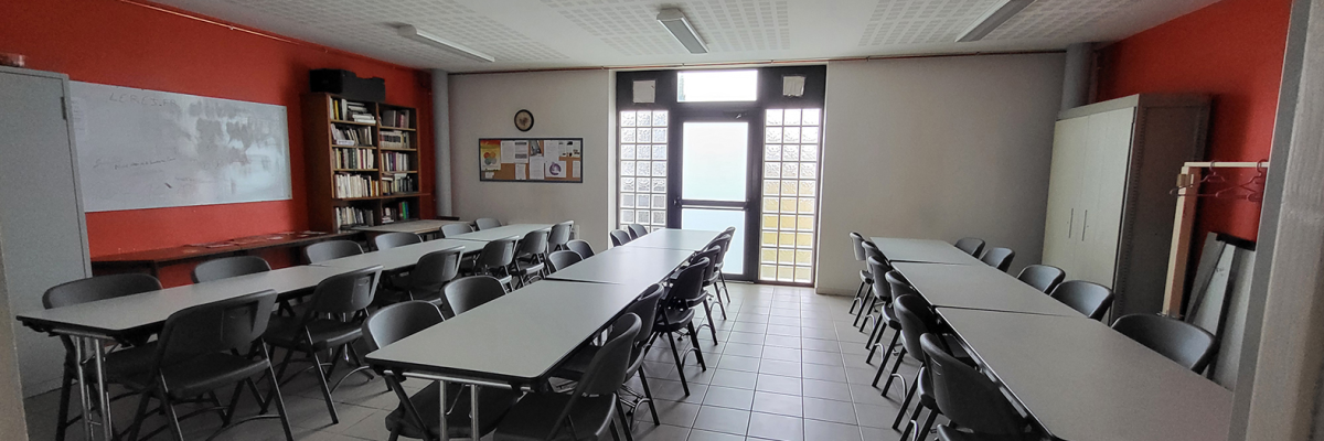 Salle avec tables, chaises, tableau blanc, étagères, porte et fenêtres.