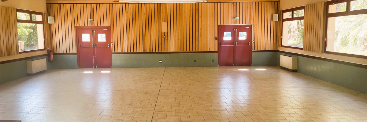 Salle vide avec sol carrelé, murs en bois, deux portes rouges, fenêtres et poutres apparentes.