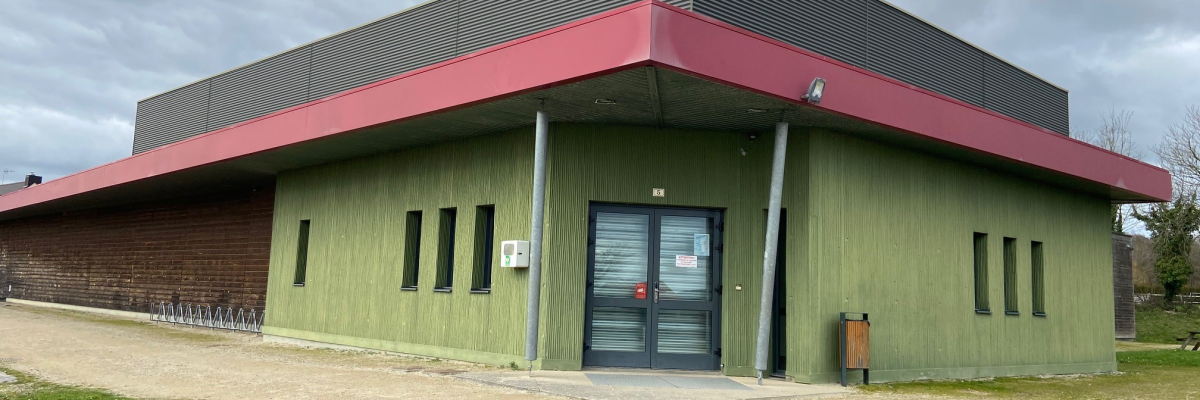 Bâtiment avec façade verte, toit gris et rouge, porte centrale, vélo à gauche, poubelle à droite.