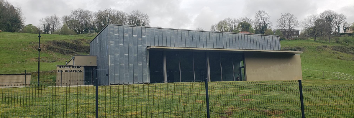 Bâtiment moderne entouré d'une clôture métallique, situé dans un champ avec des collines et des arbres en arrière-plan.