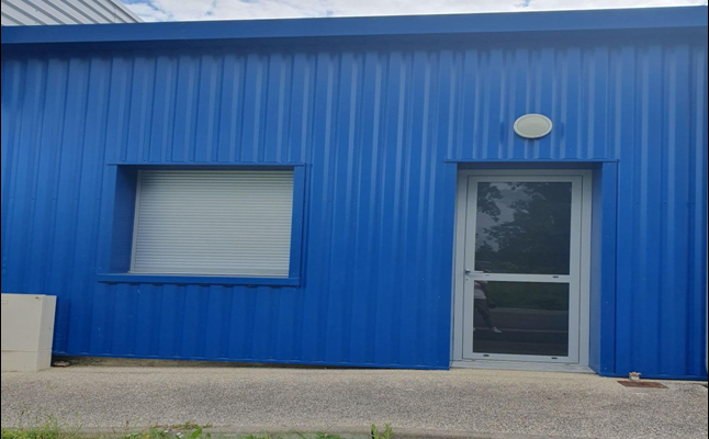 Bâtiment bleu avec porte vitrée, fenêtre à volets roulants et plaque ovale.