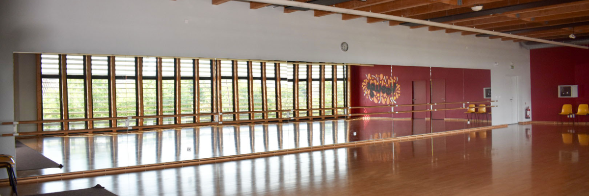 Salle de danse avec miroir, barre, tapis, chaises. Plafond en bois, grandes fenêtres.