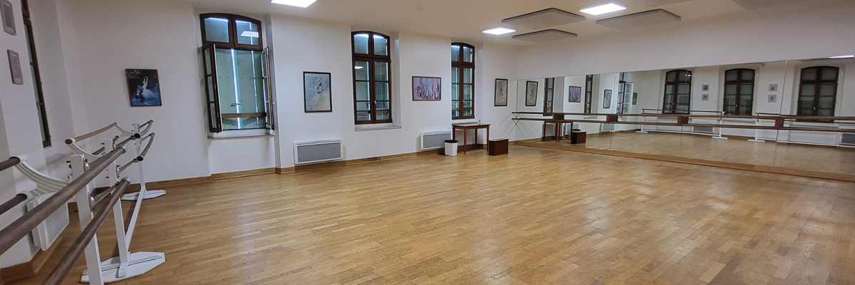 Salle de danse avec parquet, barre de ballet, fenêtres et tableaux.