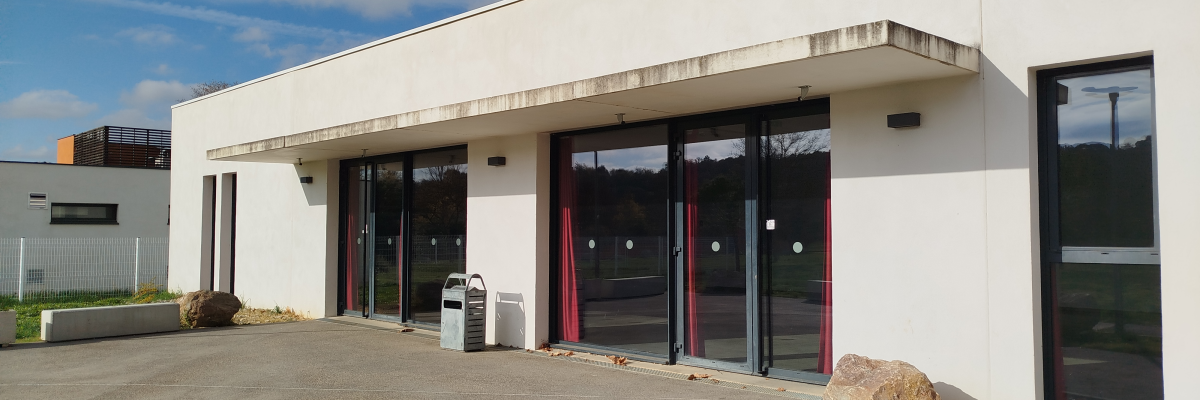 Bâtiment blanc avec grandes baies vitrées et portes, parking et rochers.