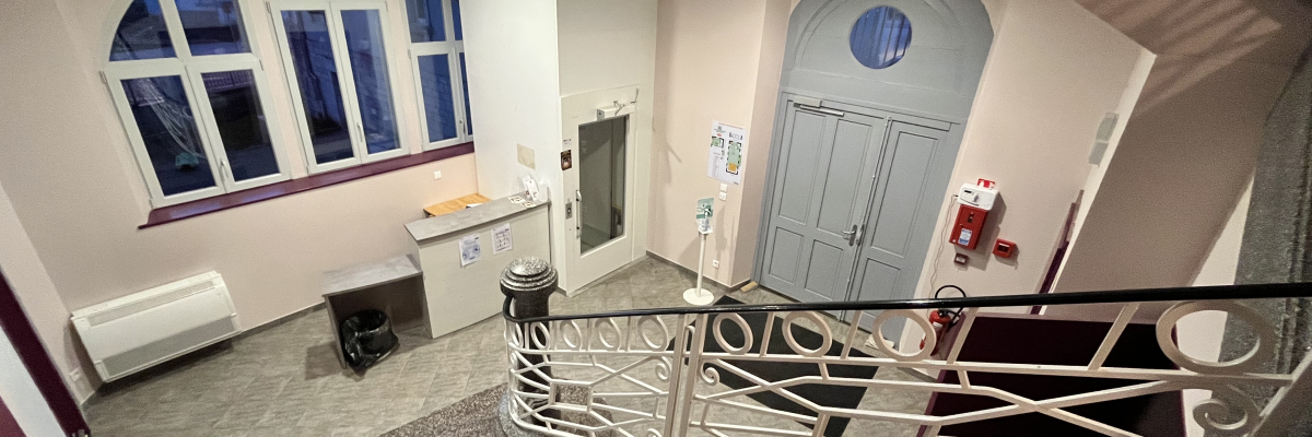 Palier avec escalier, fenêtres, radiateur, porte, rampe, extincteur et tableau d'affichage.