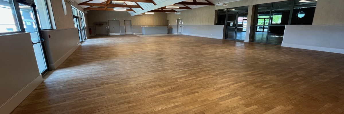 Grande salle avec parquet, poutres apparentes, luminaires suspendus, portes vitrées et fenêtres.