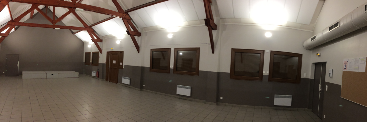 Salle avec plafond haut, poutrelles apparentes, fenêtres, portes, radiateurs, interrupteurs.