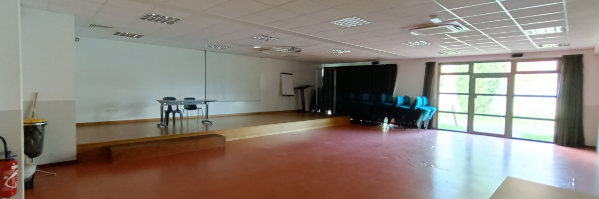Salle vide avec tables, chaises, rideau, tableau, projecteur et extincteur.