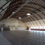 Grand espace intérieur avec toit en arc, sol en bois et quelques portes au fond.