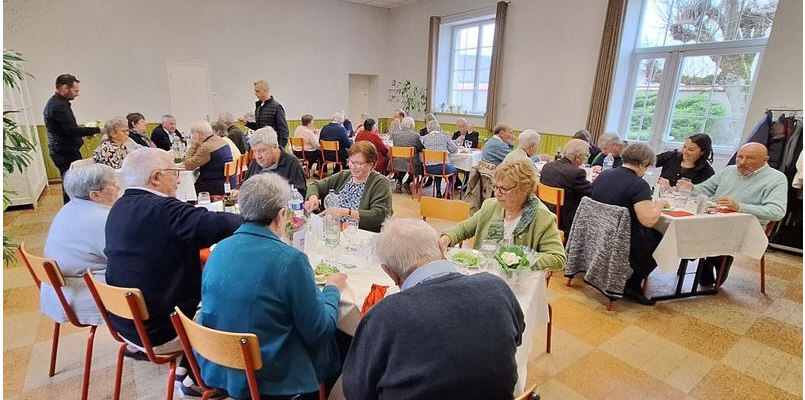 Salle avec tables, chaises, serviettes, verres et plantes. Personnes assises et debout.